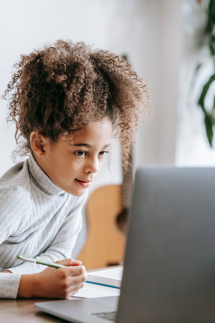 Positive African American girl with curly hair taking notes on paper while looking at screen of netbook during studies at home with guitar on blurred background