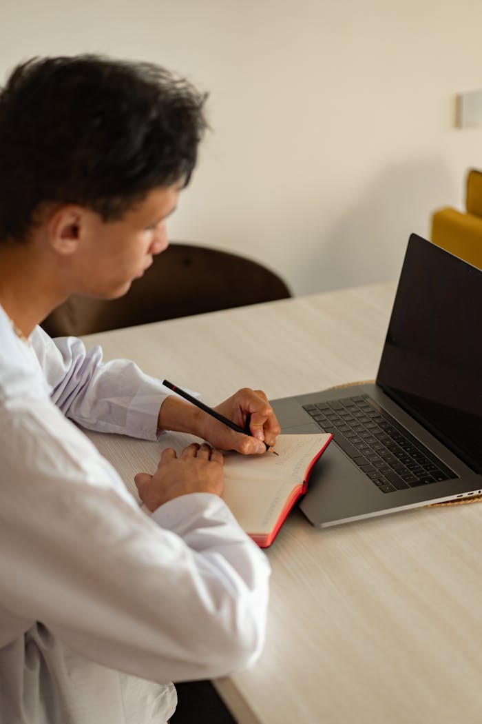 A young man focusing on his studies at home, using a laptop and notebook.